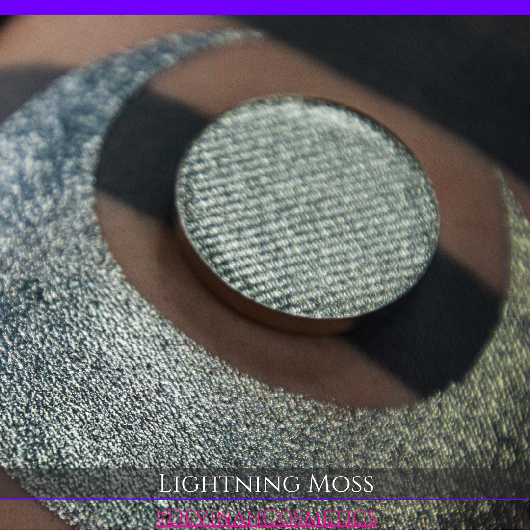 A round eyeshadow pan labeled 'LIGHTNING MOSS' made by Devinah Cosmetics, placed over a large crescent moon swatch of the same shimmering shifty eyeshadow on skin. The eyeshadow has a duochrome multichrome shift between colors pewter, silver, blue, green.