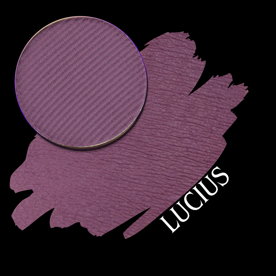 A close-up of a eyeshadow pan in a dusty mauve-purple shade, shown in a swatch over a black background with the name LUCIUS in white text.