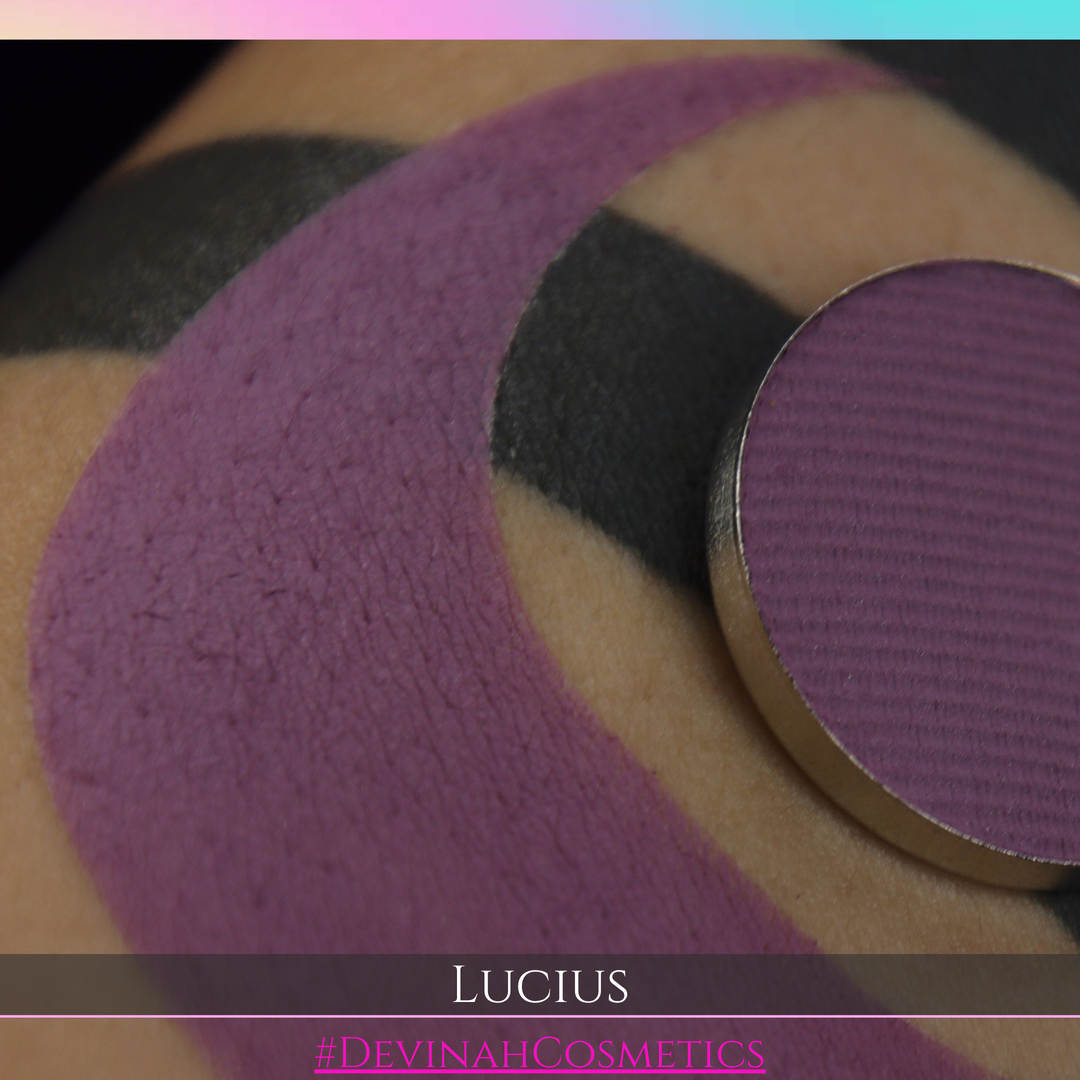 A close-up of a makeup swatch showing a large curved sweep of mauve-purple pigment on skin, with a round pan of the same shade resting on the right side. Text at the bottom reads 'LUCIUS' and '#DEVINAHCOSMETICS'.