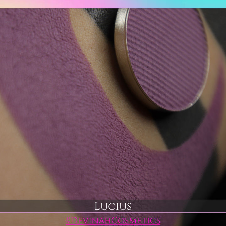 A close-up of a makeup palette showing a textured mauve-purple eyeshadow pan with a gold-toned rim, placed over a swatch of matching mauve-purple powder on beige and black cream makeup. The text 'LUCIUS' and '#DEVINAHCOSMETICS' appear at the bottom.
