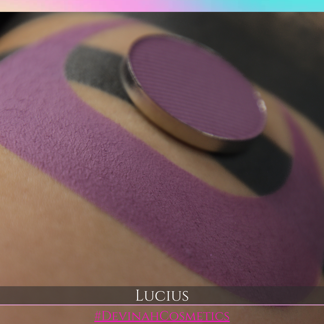 A close-up of a makeup swatch on skin showing a dusty mauve-purple color, with a round makeup pan resting on top. Text at the bottom reads 'LUCIUS' and '#DEVINAHCOSMETICS'.