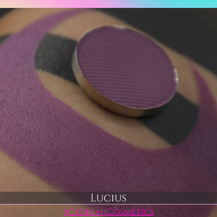 A close-up of a round makeup pan containing a deep plum purple eyeshadow, resting on top of a swatched application of the same shade on skin. The shade name 'LUCIUS' and brand '#DEVINAHCOSMETICS' are displayed in text overlays at the bottom.