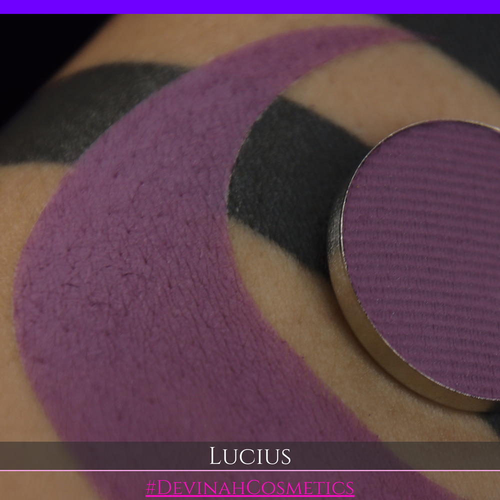 A close-up of a eyeshadow pan in a dusty mauve-purple shade, shown in a swatch over a black background with the name LUCIUS in white text made by Devinah Cosmetics.