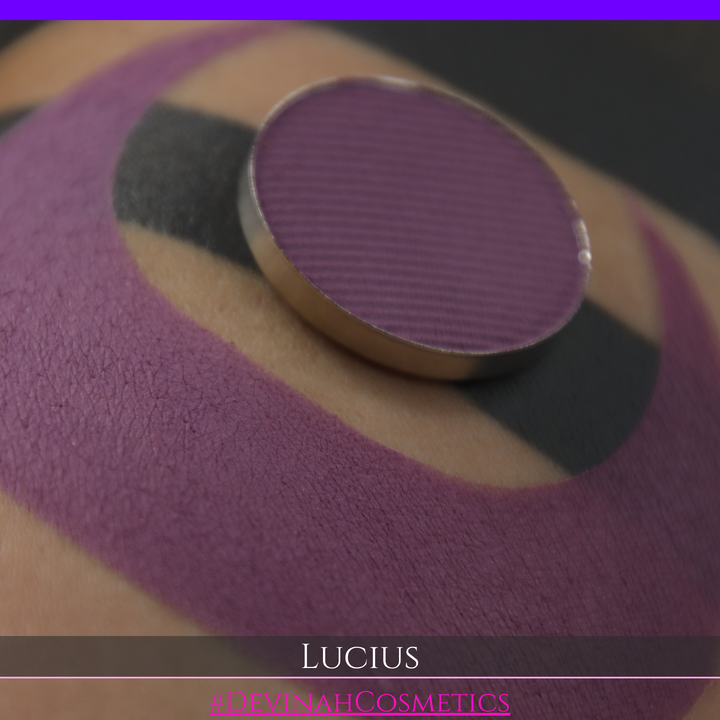 A close-up of a eyeshadow pan in a dusty mauve-purple shade, shown in a swatch over a black background with the name LUCIUS in white text made by Devinah Cosmetics.