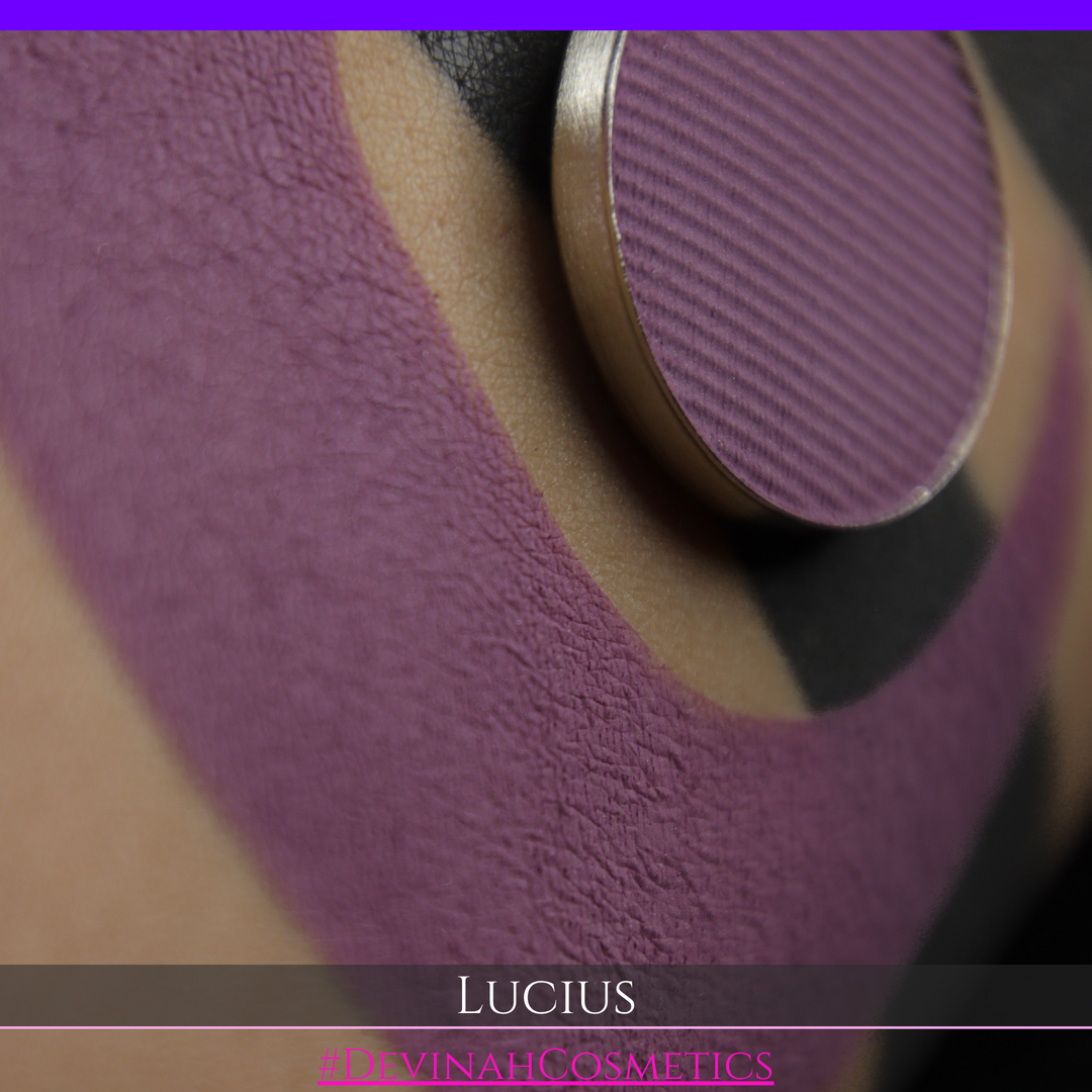 A close-up of a eyeshadow pan in a dusty mauve-purple shade, shown in a swatch over a black background with the name LUCIUS in white text made by Devinah Cosmetics.