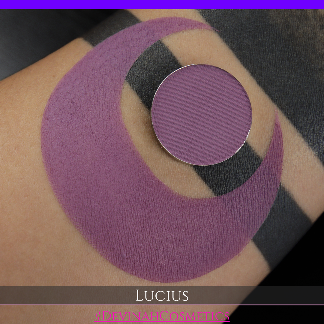 A close-up of a eyeshadow pan in a dusty mauve-purple shade, shown in a swatch over a black background with the name LUCIUS in white text made by Devinah Cosmetics.