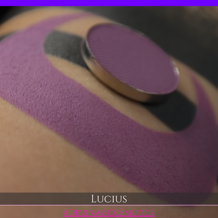 A close-up of a eyeshadow pan in a dusty mauve-purple shade, shown in a swatch over a black background with the name LUCIUS in white text made by Devinah Cosmetics.