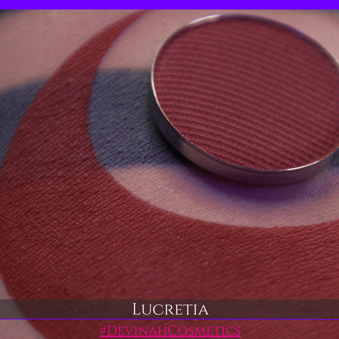 A close-up of a round eyeshadow pan in the shade 'Lucretia' by Devinah Cosmetics, resting on top of a swatched makeup base on skin. The eyeshadow is a deep, muted burgundy-red, and the background shows a swatch of the matching base on skin with text overlays at the bottom.