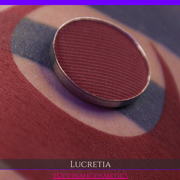 A single round eyeshadow pan in a deep rosy burgundy shade, branded 'LUCRETIA' and labeled '#DEVINAHCOSMETICS' on a dark banner at the bottom. The pan is set against a textured background of red, beige, and blue concentric circles.