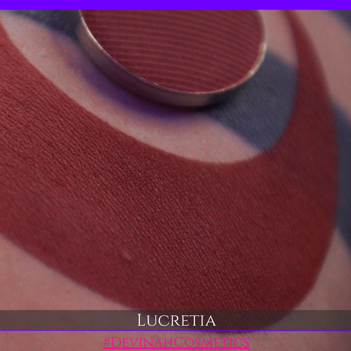 A close-up of a deep brick red or burgundy cosmetic swatch on skin, with a round metal pan resting above it. Text at the bottom reads 'LUCRETIA' and '#DEVINAHCOSMETICS'.