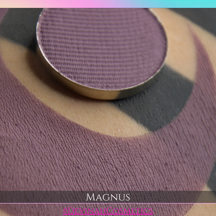 A close-up of a round makeup pan in a dusty mauve-purple shade resting on top of a matching textured swatch labeled MAGNUS.