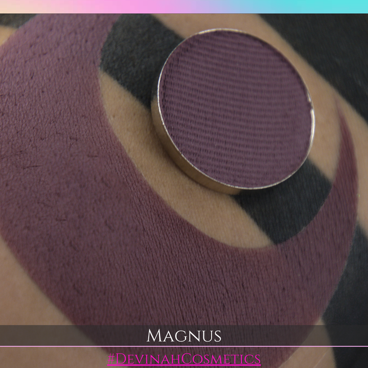 Close-up of a black makeup sponge or applicator pad with a circular metal rim, resting on skin. Text at the bottom reads 'MAGNUS' and '#DEVINAHCOSMETICS'.