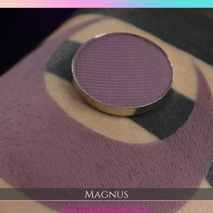A close-up of a black circular cosmetic pan or compact with a metallic rim, resting on a beige and black textured surface. Text at the bottom reads 'MAGNUS and '#DEVINAHCOSMETICS'.