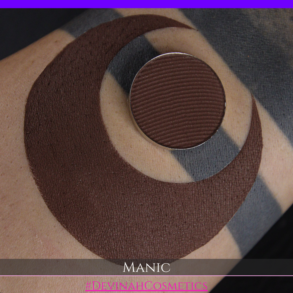 A close-up of a swatch on skin, showing a circular dark brown eyeshadow pan where the text 'MANIC' is visible at the bottom.