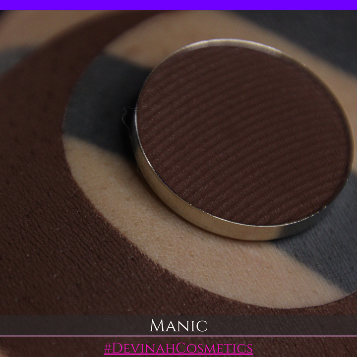 A close-up of a round pan of brown eyeshadow labeled 'Manic' at the bottom, with the shadow applied in a crescent moon on skin.