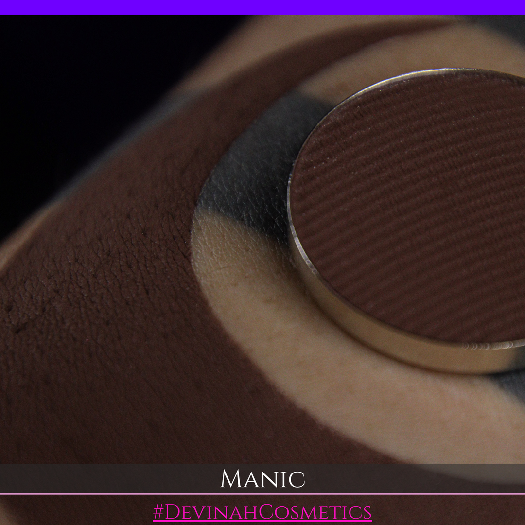 A close-up of a round makeup pan containing a dark brown shade, swatched on skin. The text 'MANIC' and '#DEVINAHCOSMETICS' are displayed at the bottom.