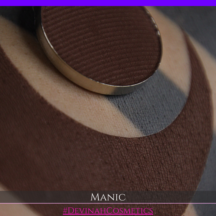 A close-up of a makeup swatch showing a dark brown eyeshadow pan swatched on skin. The text 'MANIC' and '#DEVINAHCOSMETICS' are displayed at the bottom.