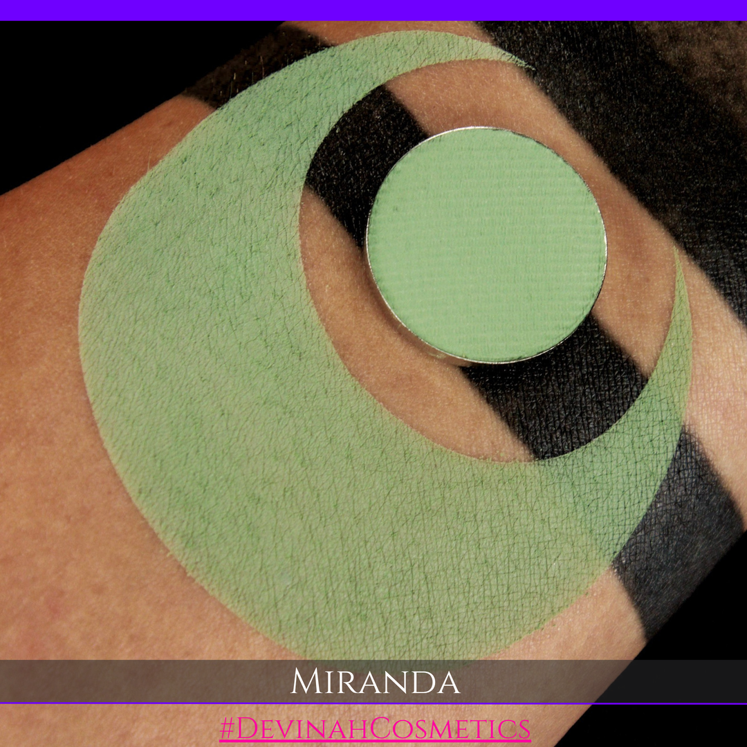 A close-up of a person's arm with a large light green makeup shape applied, alongside a matching round eyeshadow or powder pan. Text at the bottom reads 'MIRANDA' and '#DEVINACHECOSMETICS'.