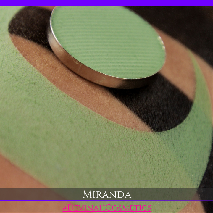 A close-up of a green textured cosmetic pan or compact lid resting on a green and black patterned surface, with the word 'MIRANDA' and a Devinah Cosmetics brand name at the bottom.