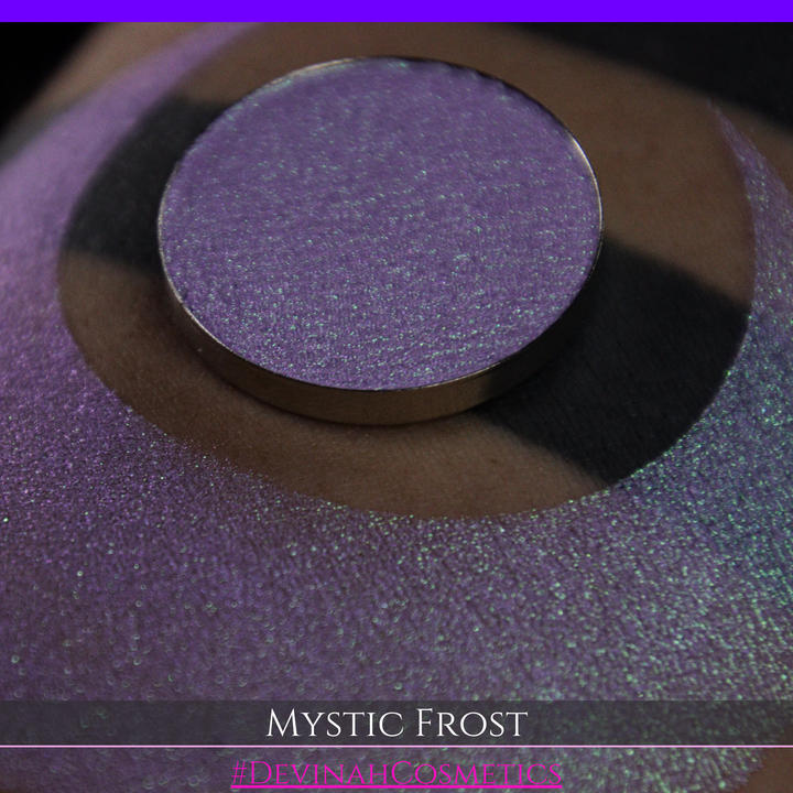 A textured eyeshadow swatch labeled 'MYSTIC FROST' with a purple-to-teal gradient, showing a circular eyeshadow makeup pan on a black background hand made by Devinah Cosmetics.