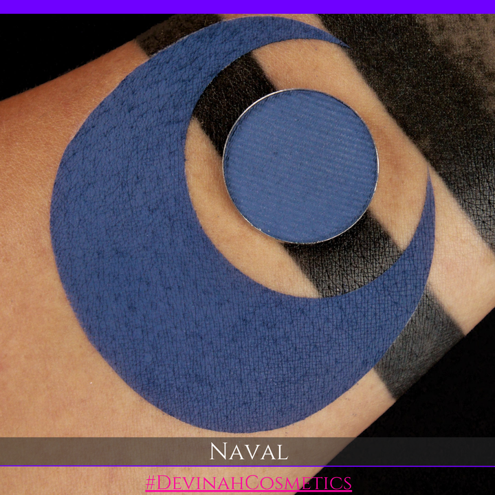 A close-up of a person's arm with a large blue crescent moon makeup smudge, alongside a round pan of blue eyeshadow labeled 'NAVAL' by Devinah Cosmetics.
