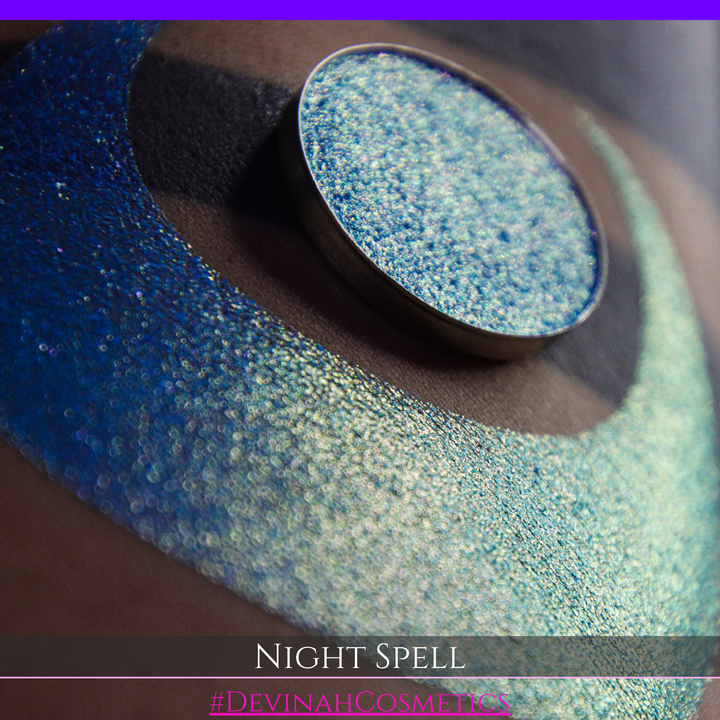 A close-up of a person's arm with a large crescent moon makeup swatch and circular makeup eyeshadow pan in a shimmery blue-to-purple gradient, labeled 'Night Spell' with '#DevinahCosmetics' text at the bottom.