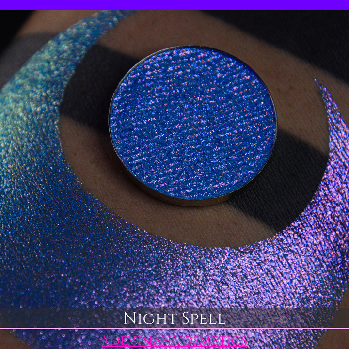 A close-up of a person's arm with a large crescent moon makeup swatch and circular makeup eyeshadow pan in a shimmery blue-to-purple gradient, labeled 'Night Spell' with '#DevinahCosmetics' text at the bottom.
