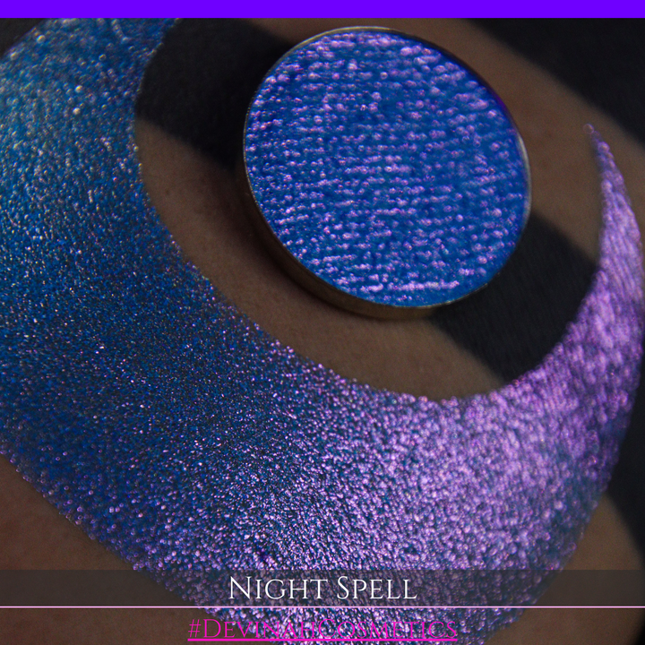 A close-up of a person's arm with a large crescent moon makeup swatch and circular makeup eyeshadow pan in a shimmery blue-to-purple gradient, labeled 'Night Spell' with '#DevinahCosmetics' text at the bottom.