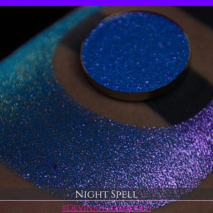 A close-up of a person's arm with a large crescent moon makeup swatch and circular makeup eyeshadow pan in a shimmery blue-to-purple gradient, labeled 'Night Spell' with '#DevinahCosmetics' text at the bottom.