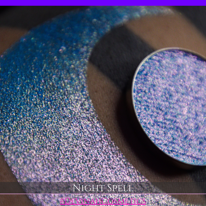 A close-up of a person's arm with a large crescent moon makeup swatch and circular makeup eyeshadow pan in a shimmery blue-to-purple gradient, labeled 'Night Spell' with '#DevinahCosmetics' text at the bottom.