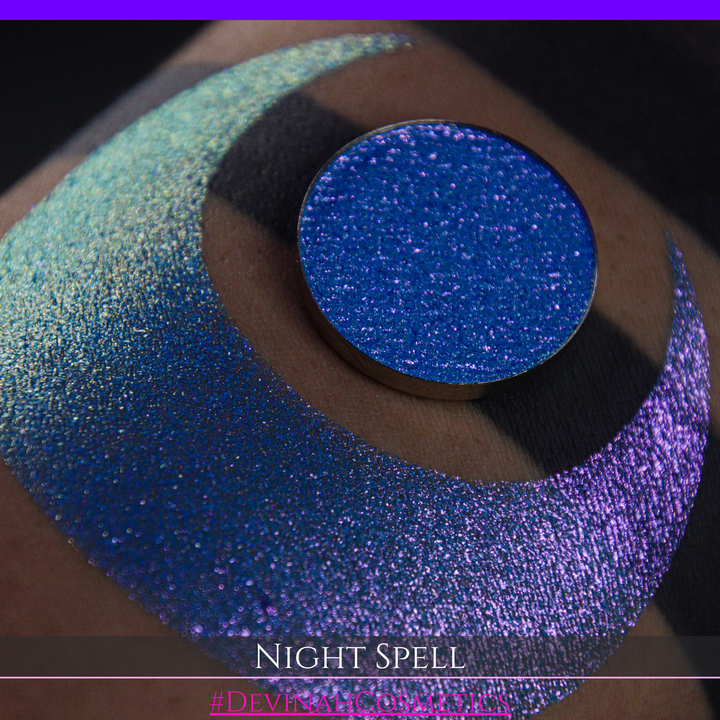 A close-up of a person's arm with a large crescent moon makeup swatch and circular makeup eyeshadow pan in a shimmery blue-to-purple gradient, labeled 'Night Spell' with '#DevinahCosmetics' text at the bottom.