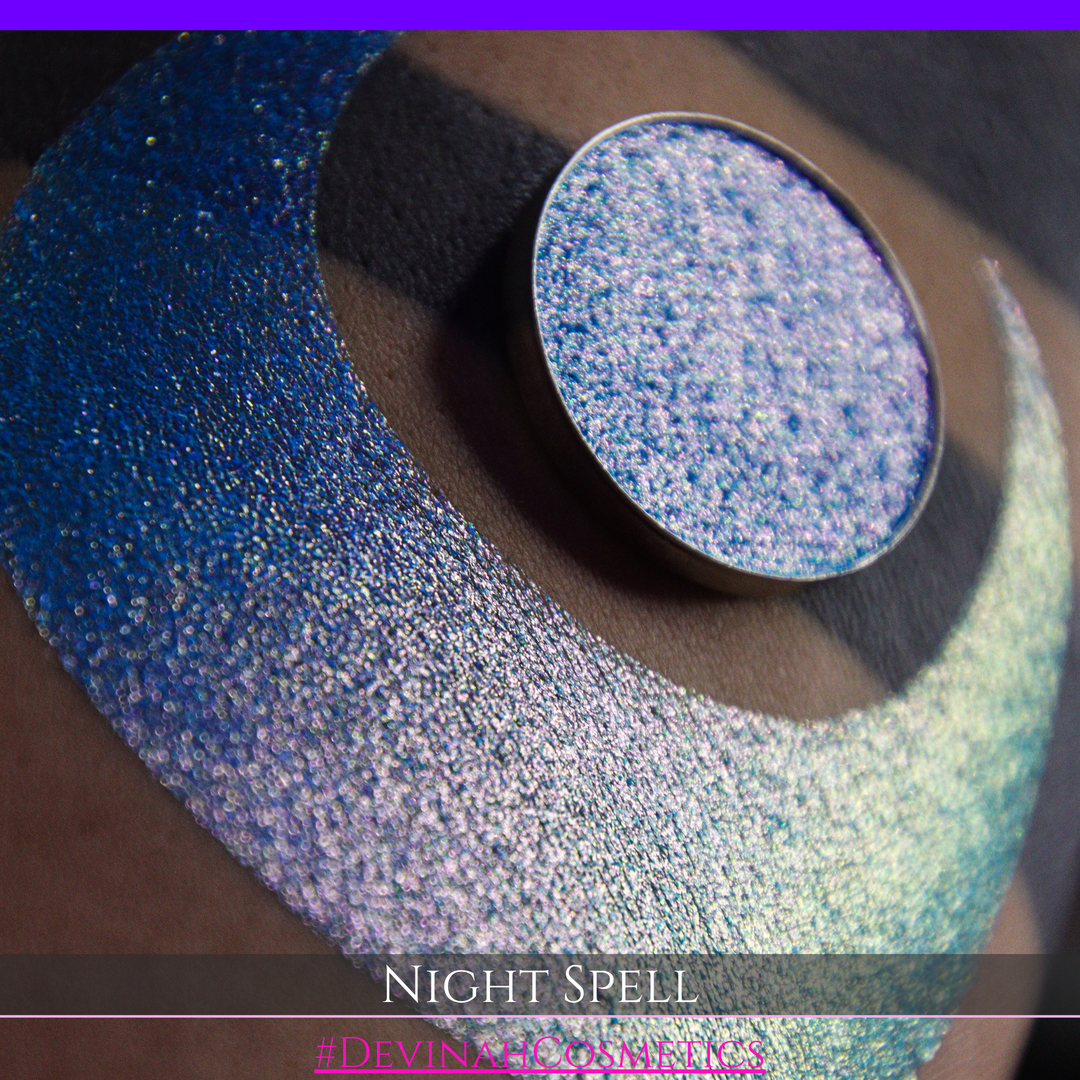 A close-up of a person's arm with a large crescent moon makeup swatch and circular makeup eyeshadow pan in a shimmery blue-to-purple gradient, labeled 'Night Spell' with '#DevinahCosmetics' text at the bottom.