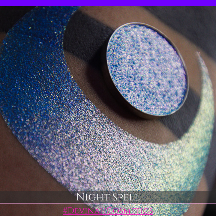 A close-up of a person's arm with a large crescent moon makeup swatch and circular makeup eyeshadow pan in a shimmery blue-to-purple gradient, labeled 'Night Spell' with '#DevinahCosmetics' text at the bottom.