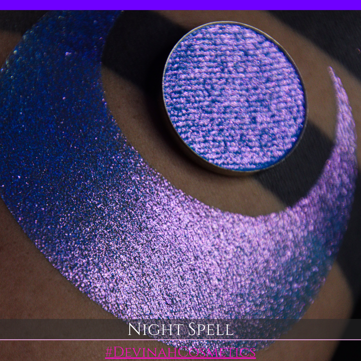 A close-up of a person's arm with a large crescent moon makeup swatch and circular makeup eyeshadow pan in a shimmery blue-to-purple gradient, labeled 'Night Spell' with '#DevinahCosmetics' text at the bottom.