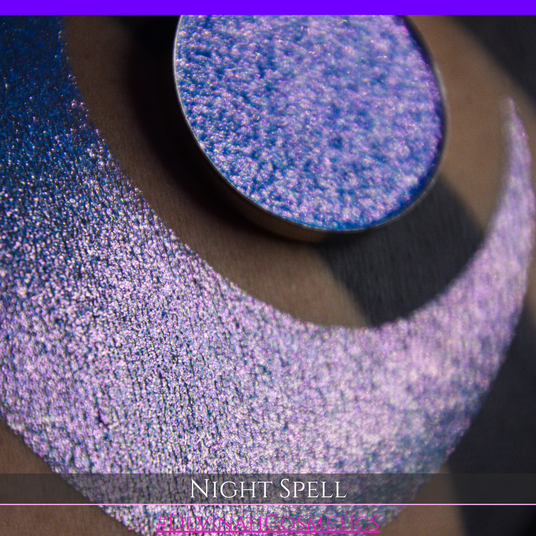 A close-up of a person's arm with a large crescent moon makeup swatch and circular makeup eyeshadow pan in a shimmery blue-to-purple gradient, labeled 'Night Spell' with '#DevinahCosmetics' text at the bottom.