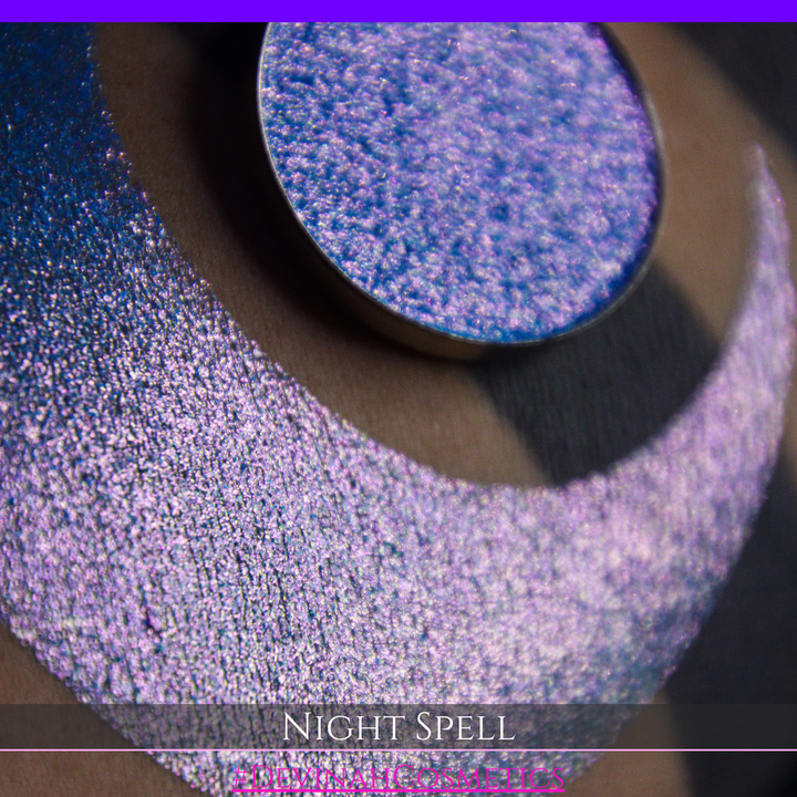 A close-up of a person's arm with a large crescent moon makeup swatch and circular makeup eyeshadow pan in a shimmery blue-to-purple gradient, labeled 'Night Spell' with '#DevinahCosmetics' text at the bottom.