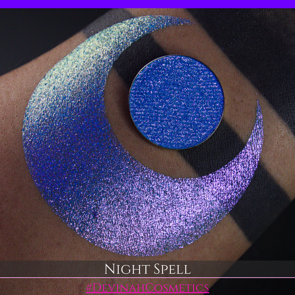 A close-up of a person's arm with a large crescent moon makeup swatch and circular makeup eyeshadow pan in a shimmery blue-to-purple gradient, labeled 'Night Spell' with '#DevinahCosmetics' text at the bottom.