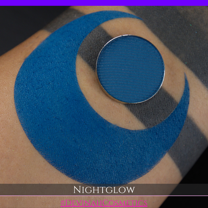 A close-up of a person's arm with a large blue crescent-shaped makeup swatch and a circular blue eyeshadow pan placed on top. Text at the bottom reads 'NIGHTGLOW' and a pink handle '@DEVINAHCOSMETICS'.