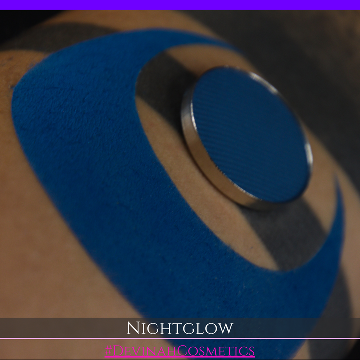 A close-up of a blue circular cosmetic eyeshadow pan resting on a tan arm with a large blue crescent swatch. The text 'NIGHTGLOW' and '#DEVINAHCOSMETICS' appears at the bottom.