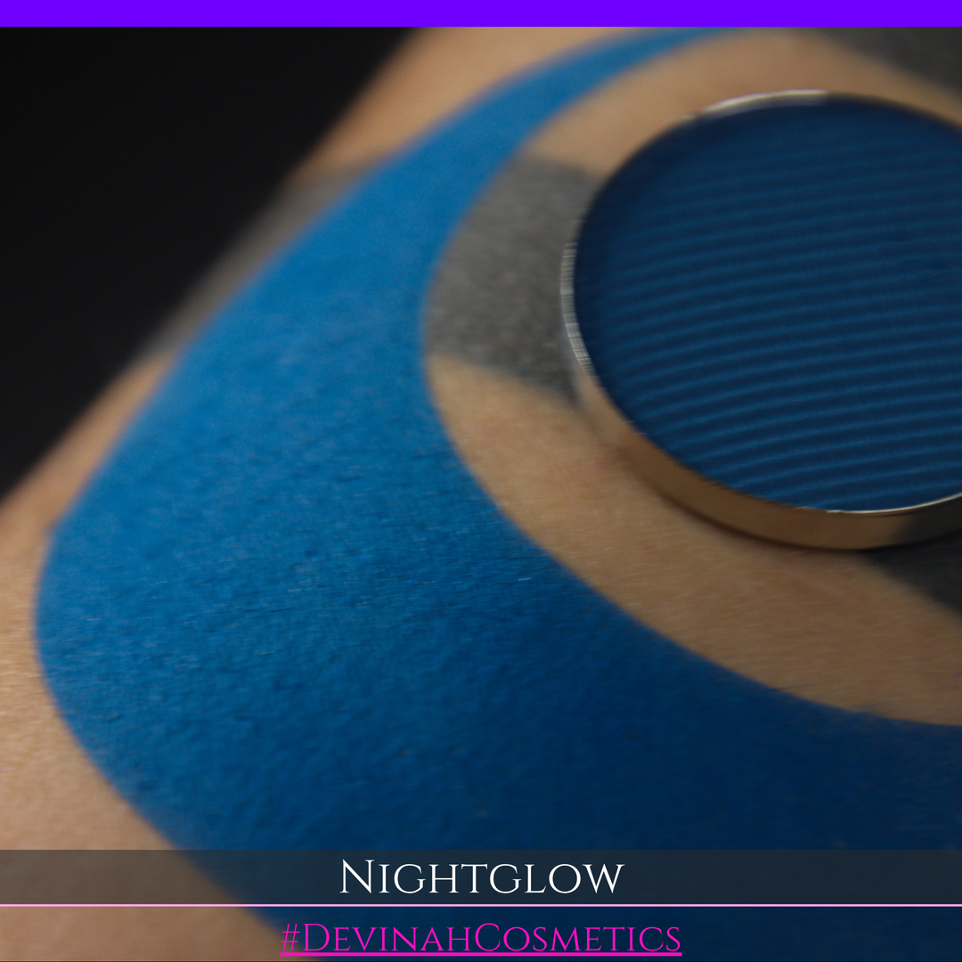 A close-up of a blue eyeshadow pan resting on a person's upper arm, with the shade name 'NIGHTGLOW' and the brand 'DEVINAH COSMETICS' shown in text overlays at the bottom.
