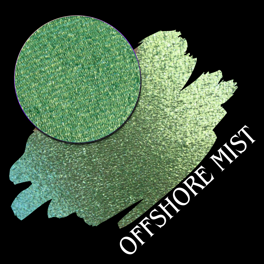 A round eyeshadow pan labeled 'OFFSHORE MIST' made by Devinah Cosmetics, placed over a large crescent moon swatch of the same shimmering shifty eyeshadow on skin. The eyeshadow has a duochrome multichrome shift between colors lime, green, yellow, blue. 