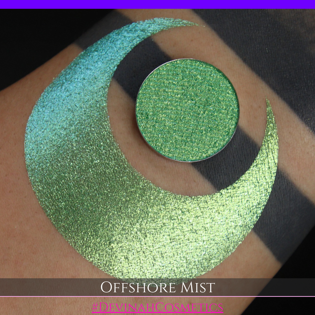 A round eyeshadow pan labeled 'OFFSHORE MIST' made by Devinah Cosmetics, placed over a large crescent moon swatch of the same shimmering shifty eyeshadow on skin. The eyeshadow has a duochrome multichrome shift between colors lime, green, yellow, blue. 