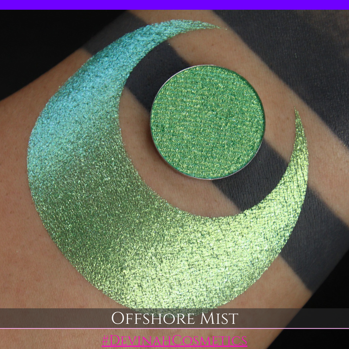 A round eyeshadow pan labeled 'OFFSHORE MIST' made by Devinah Cosmetics, placed over a large crescent moon swatch of the same shimmering shifty eyeshadow on skin. The eyeshadow has a duochrome multichrome shift between colors lime, green, yellow, blue. 