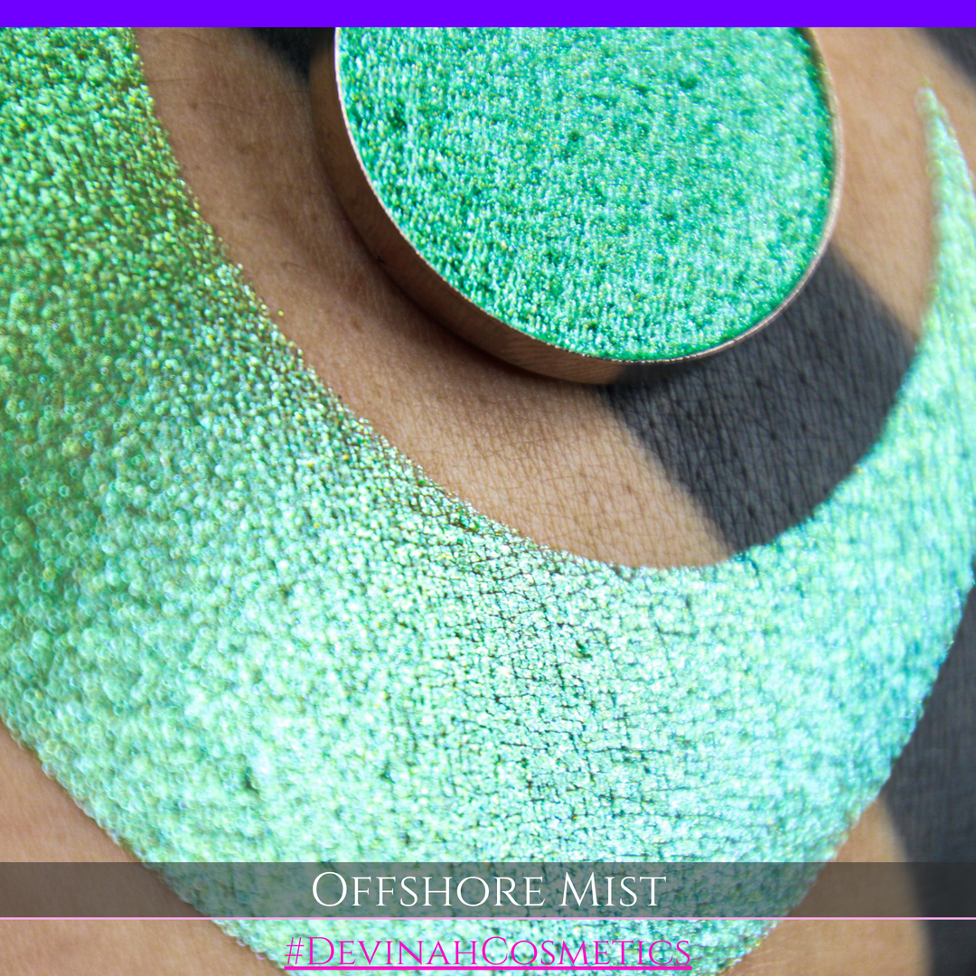 A round eyeshadow pan labeled 'OFFSHORE MIST' made by Devinah Cosmetics, placed over a large crescent moon swatch of the same shimmering shifty eyeshadow on skin. The eyeshadow has a duochrome multichrome shift between colors lime, green, yellow, blue. 