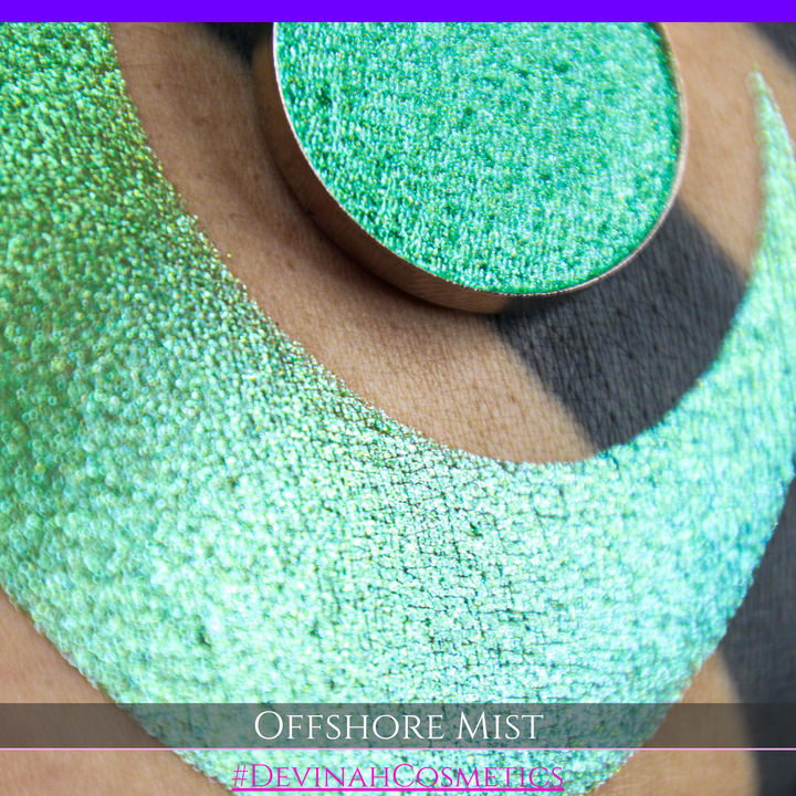 A round eyeshadow pan labeled 'OFFSHORE MIST' made by Devinah Cosmetics, placed over a large crescent moon swatch of the same shimmering shifty eyeshadow on skin. The eyeshadow has a duochrome multichrome shift between colors lime, green, yellow, blue. 
