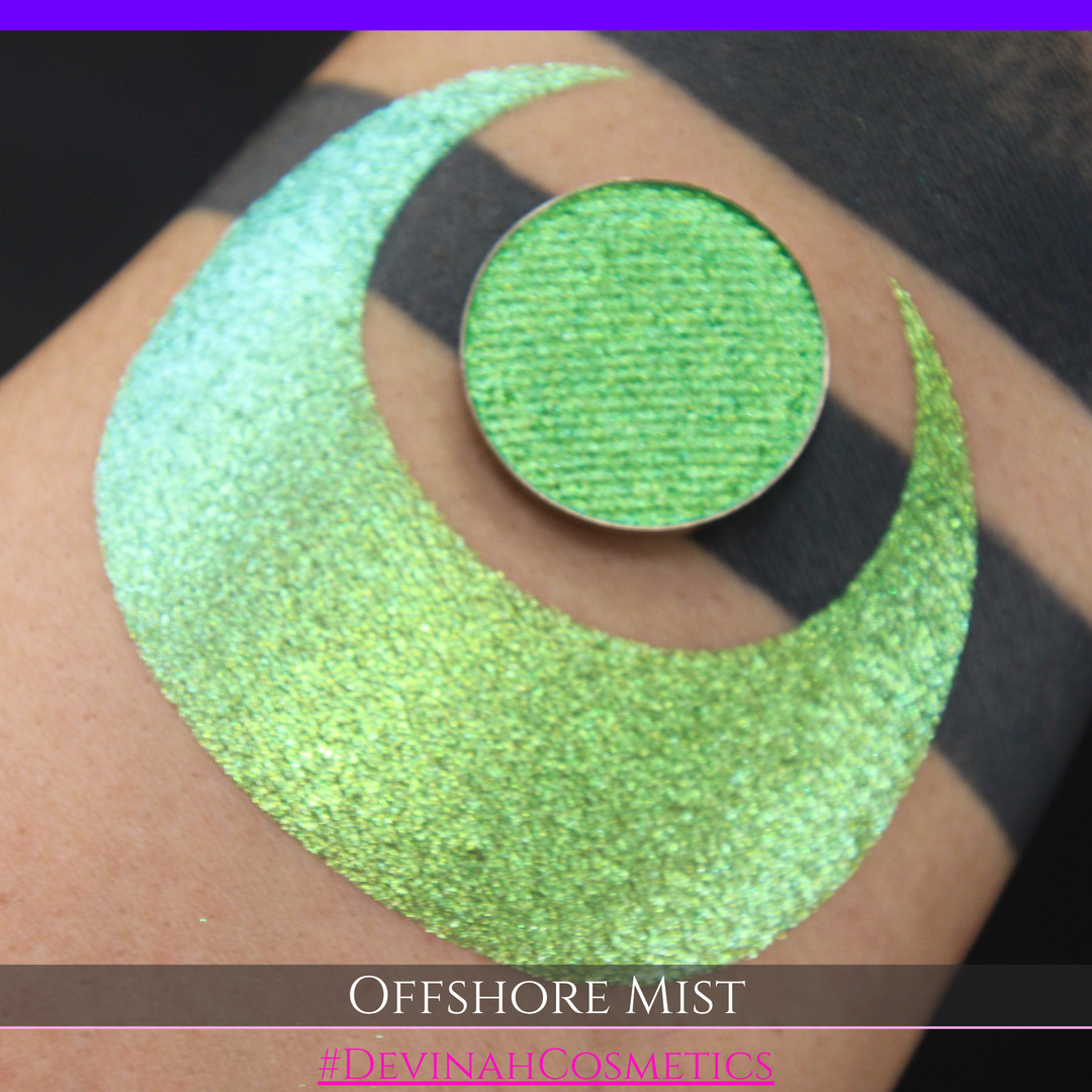 A round eyeshadow pan labeled 'OFFSHORE MIST' made by Devinah Cosmetics, placed over a large crescent moon swatch of the same shimmering shifty eyeshadow on skin. The eyeshadow has a duochrome multichrome shift between colors lime, green, yellow, blue. 