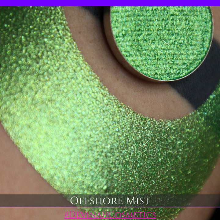 A round eyeshadow pan labeled 'OFFSHORE MIST' made by Devinah Cosmetics, placed over a large crescent moon swatch of the same shimmering shifty eyeshadow on skin. The eyeshadow has a duochrome multichrome shift between colors lime, green, yellow, blue. 