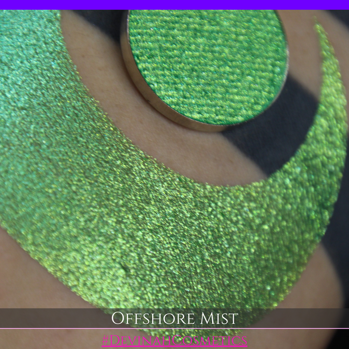 A round eyeshadow pan labeled 'OFFSHORE MIST' made by Devinah Cosmetics, placed over a large crescent moon swatch of the same shimmering shifty eyeshadow on skin. The eyeshadow has a duochrome multichrome shift between colors lime, green, yellow, blue. 