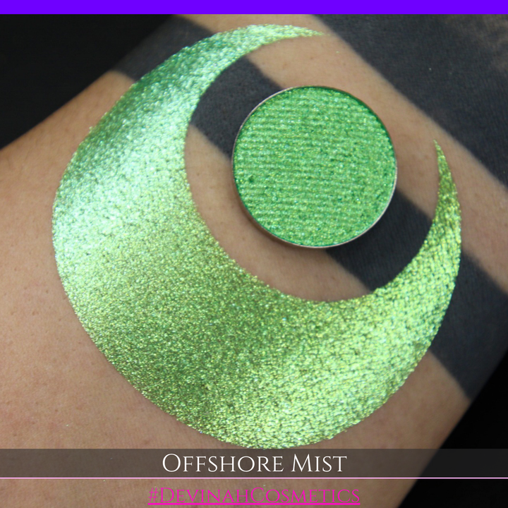 A round eyeshadow pan labeled 'OFFSHORE MIST' made by Devinah Cosmetics, placed over a large crescent moon swatch of the same shimmering shifty eyeshadow on skin. The eyeshadow has a duochrome multichrome shift between colors lime, green, yellow, blue. 