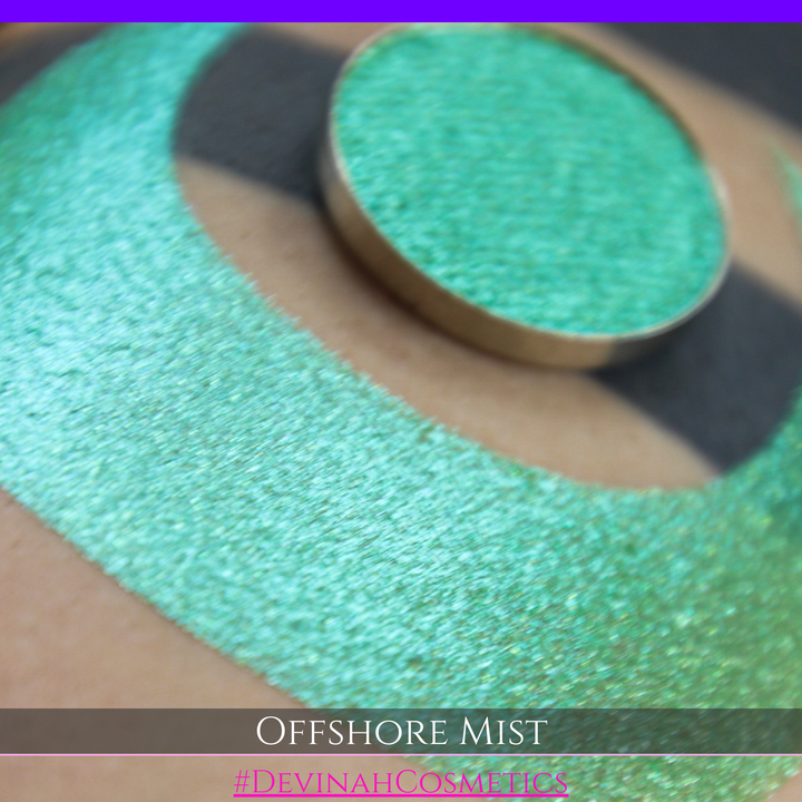A round eyeshadow pan labeled 'OFFSHORE MIST' made by Devinah Cosmetics, placed over a large crescent moon swatch of the same shimmering shifty eyeshadow on skin. The eyeshadow has a duochrome multichrome shift between colors lime, green, yellow, blue. 